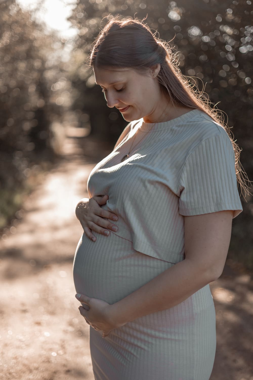 Gravid kvinde holder om maven og kigger ned i modlys på en skovvej